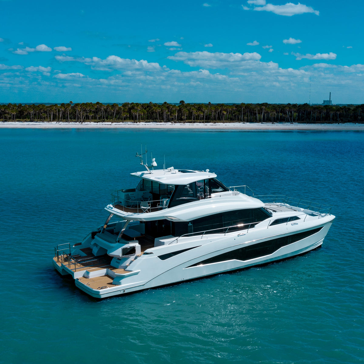 A luxury motor yacht with a white hull and dark gray accents floats on clear turquoise water, with a sandy beach, dense green palm forest, and blue sky with scattered clouds in the background.